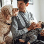 Father sitting on a couch feeding a baby with a bottle while a friendly dog looks on lovingly.